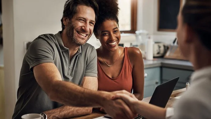Junges Paar sitzt lächelnd an einem Schreibtisch, er schüttelt die Hand eines Beraters gegenüber